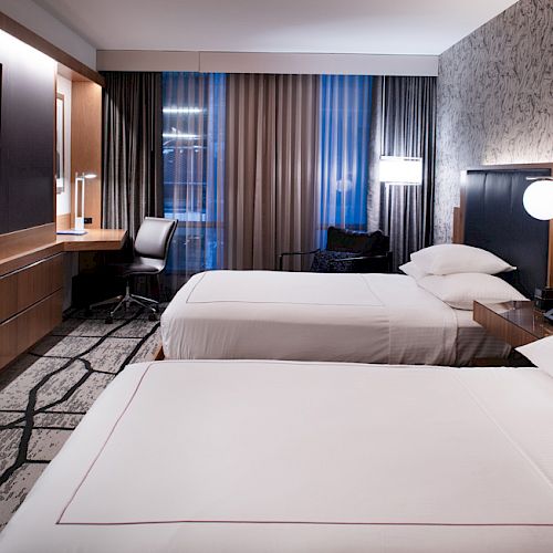 The image shows a hotel room with two beds, a flat-screen TV, a desk, and large window with curtains, creating a modern and cozy ambiance.