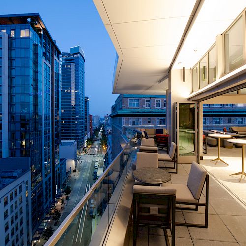 A modern rooftop patio with seating overlooks a cityscape at dusk, featuring illuminated tall buildings and street below.