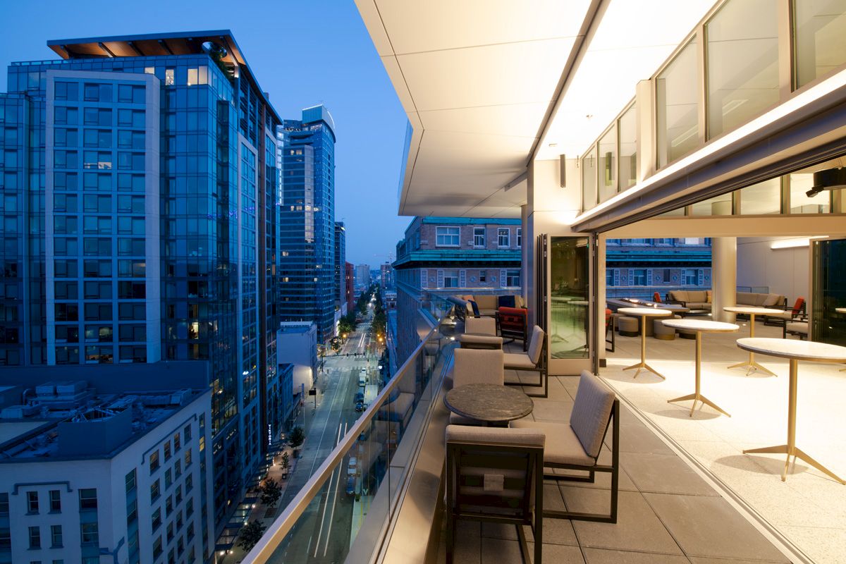 A modern cityscape at dusk with tall buildings and a stylish balcony featuring tables and chairs, overlooking the illuminated street.