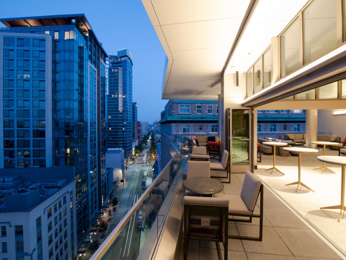 A modern cityscape at dusk with tall buildings and a stylish balcony featuring tables and chairs, overlooking the illuminated street.