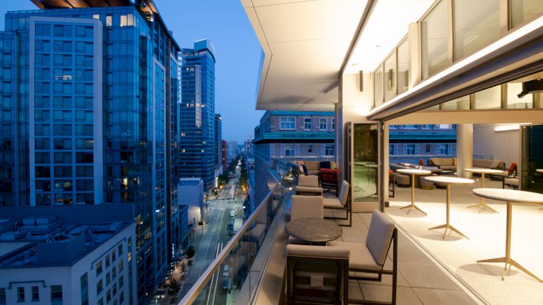 A modern cityscape at dusk with tall buildings and a stylish balcony featuring tables and chairs, overlooking the illuminated street.