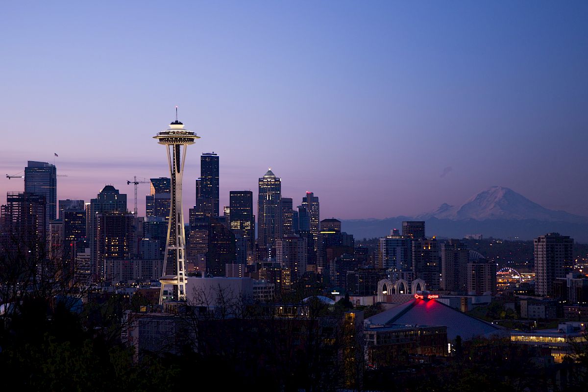 Skyline of Seattle at dusk with the Space Needle and city buildings, Mount Rainier in the background, under a clear sky.