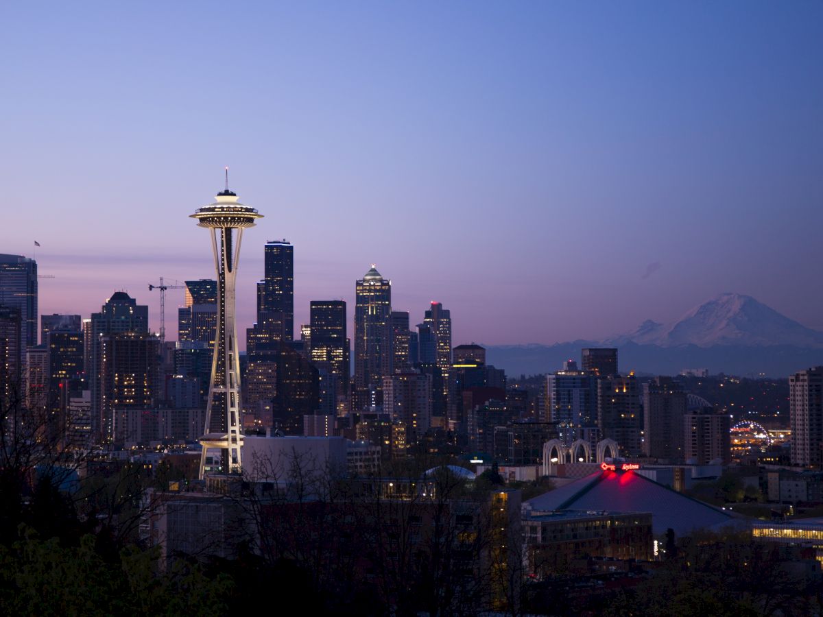 Skyline of Seattle at dusk with the Space Needle and city buildings, Mount Rainier in the background, under a clear sky.
