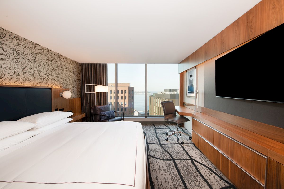 The image shows a modern hotel room with a bed, large TV, desk, chair, and a window offering a view of city buildings.