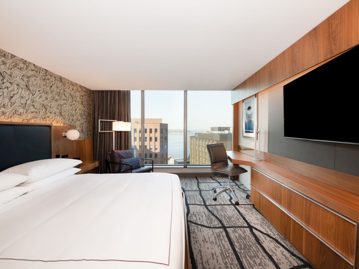 The image shows a modern hotel room with a bed, large TV, desk, chair, and a window offering a view of city buildings.