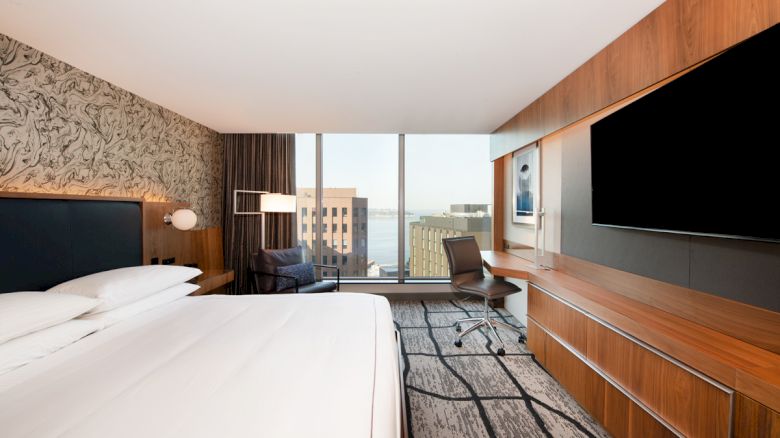 The image shows a modern hotel room with a bed, large TV, desk, chair, and a window offering a view of city buildings.