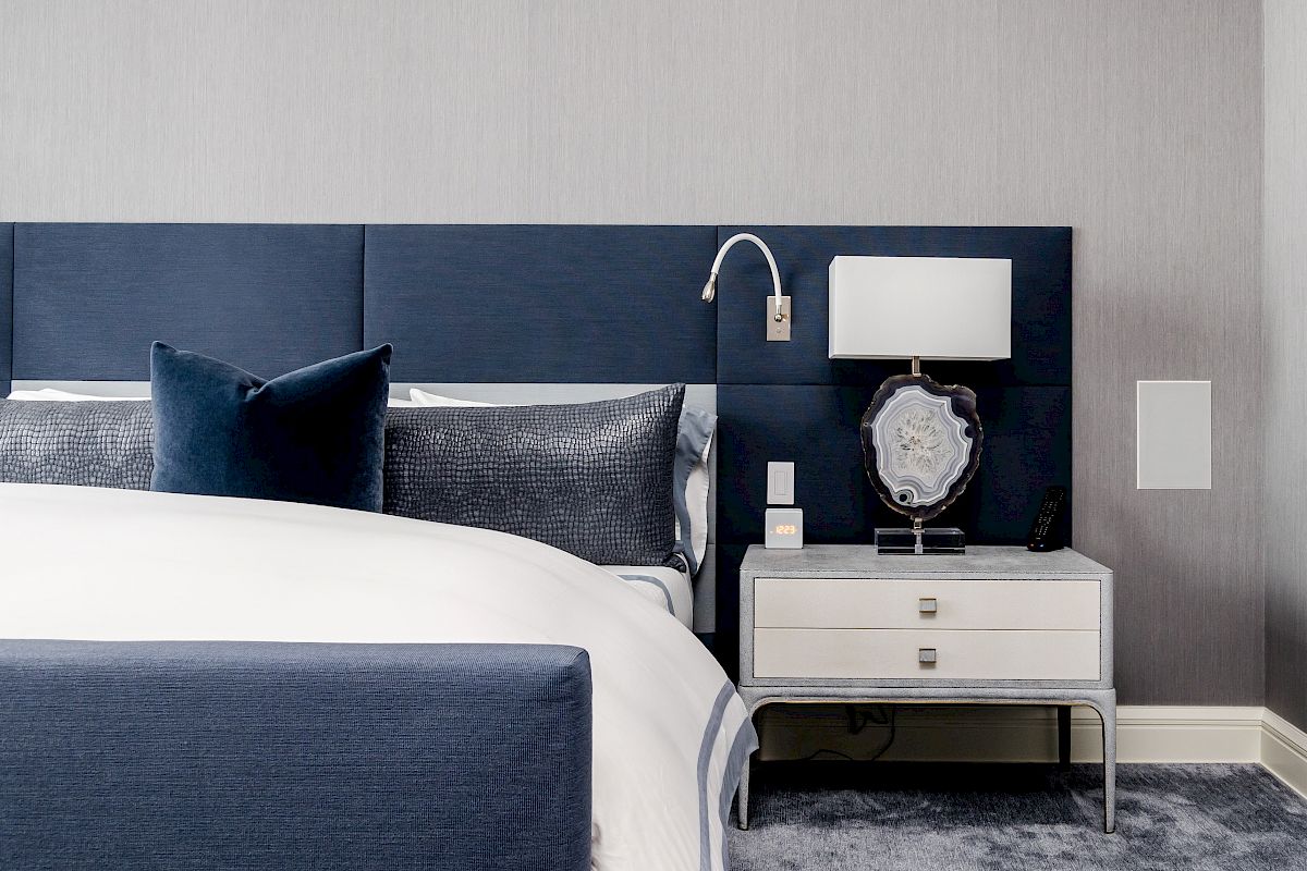 A modern bedroom with a navy headboard, white bedding, and a nightstand featuring a lamp and decorative mirror.