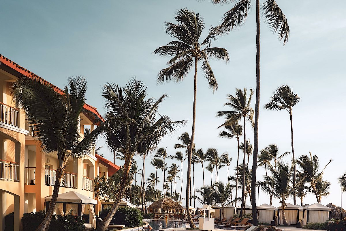 A resort pool area with lounge chairs, palm trees, and a building with balconies, set against a clear sky, creating a tropical ambiance.