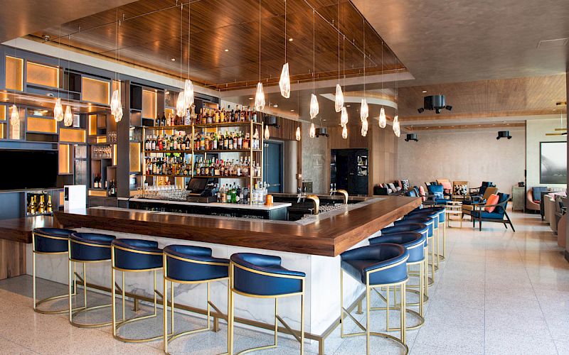 The image shows a modern bar with blue cushioned stools, a well-stocked counter, hanging lights, and an open seating area for guests.