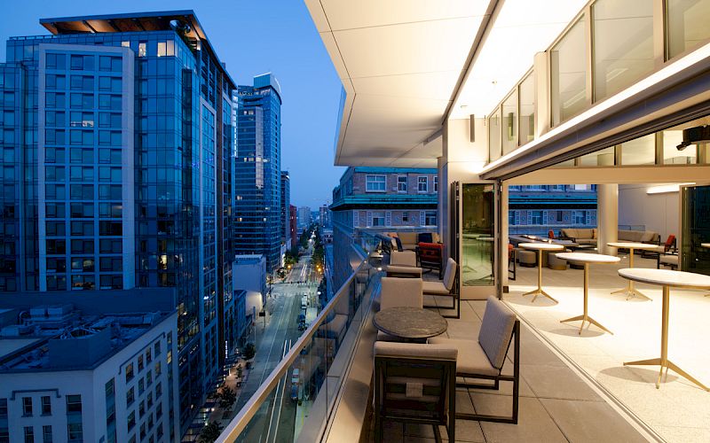 A modern balcony at dusk overlooking a city street, with tables, chairs, and tall buildings in the background.