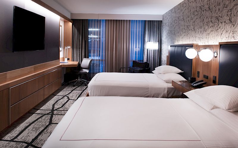 The image shows a modern hotel room with two beds, a large TV, a desk, lamps, and stylish decor in neutral tones.