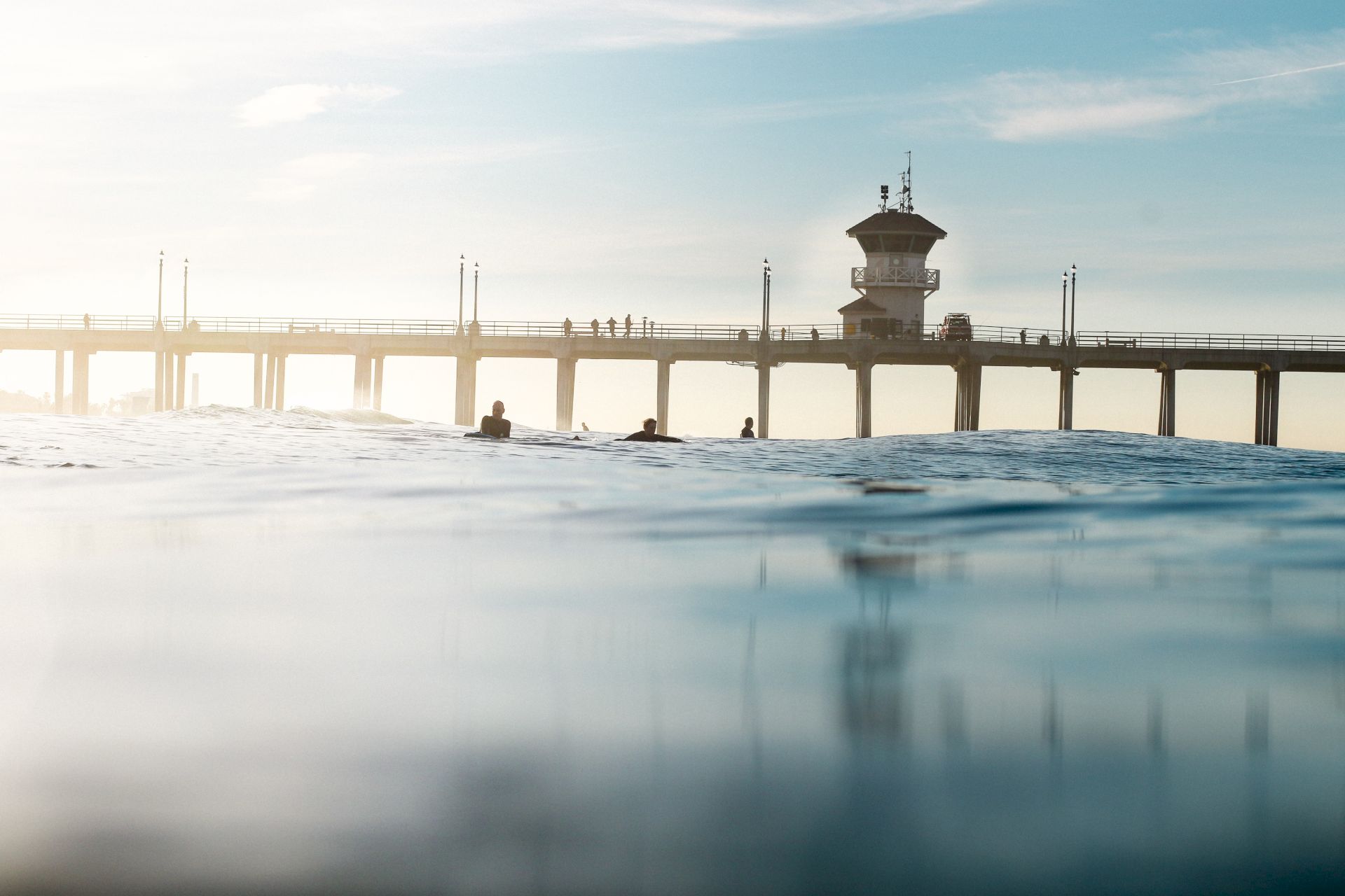 A pier extends over the ocean, with people walking atop it, under a blue sky. The water in the foreground gives a serene vibe.